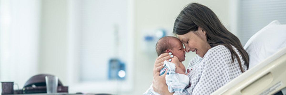A mother in hospital gown sits in hospital bed nuzzling her new baby